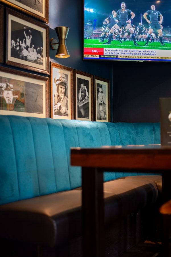 A screen showing rugby above a teal velvet booth, with framed pictures on the wall