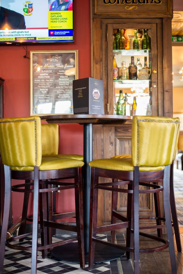 Two mustard high stools at a tall table with a menu stand, in front of a glass-fronted Whelan's cabinet of old bottles
