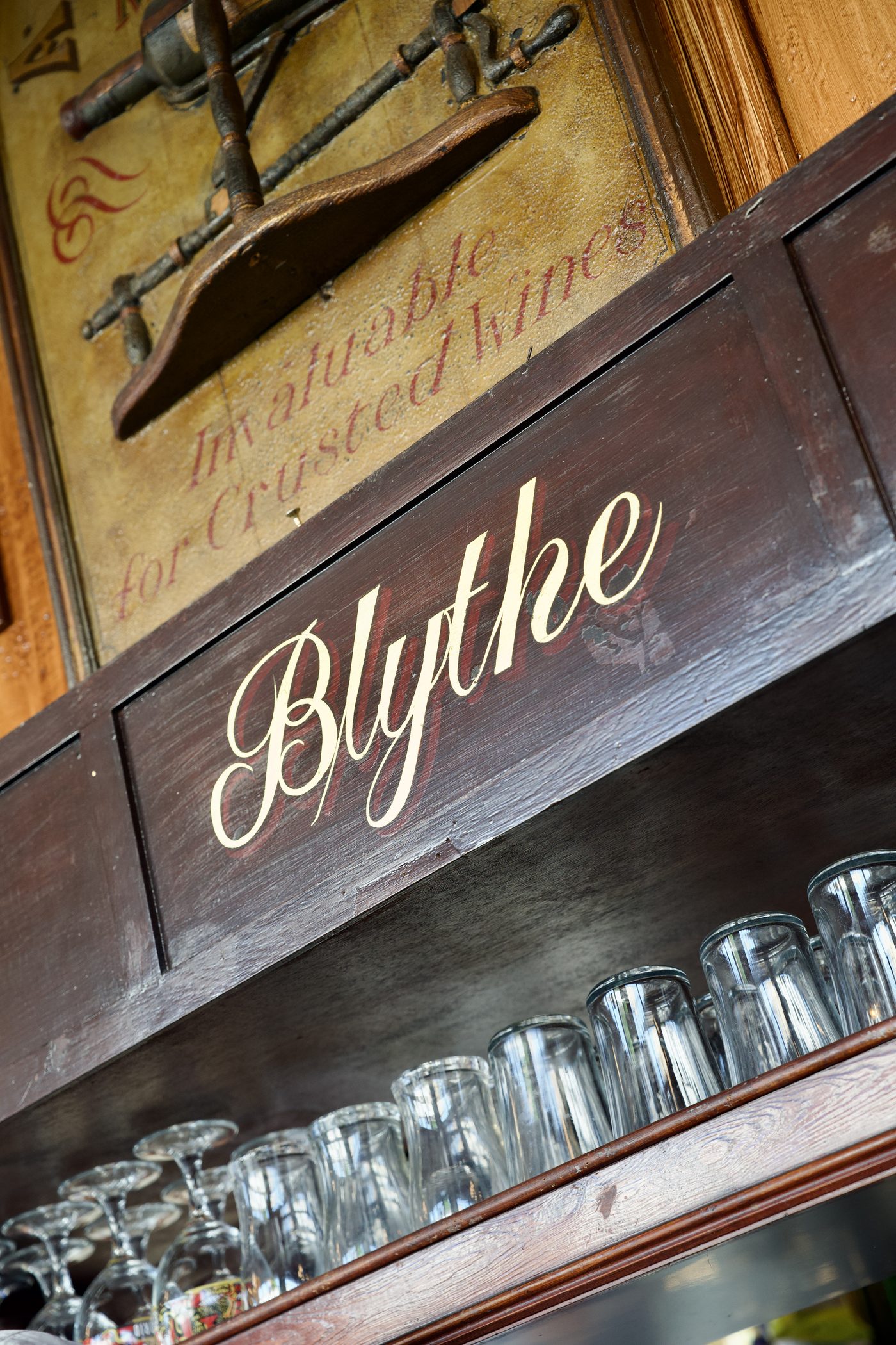 Blythe painted in cream script on the dark gantry panelling, an old corkscrew advertisement above and glasses on the shelf below