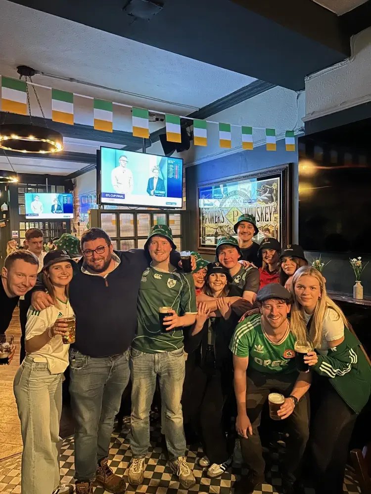 A group of Ireland fans celebrating together, green bunting overhead and a screen showing rugby coverage