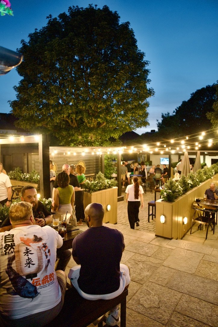 The beer garden in the evening, busy with drinkers under string lights