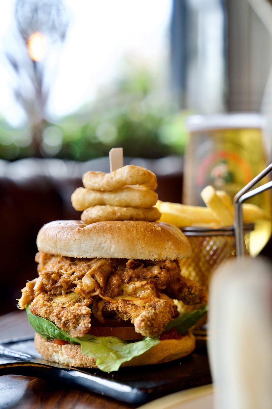 A fried chicken burger stacked with onion rings, fries alongside