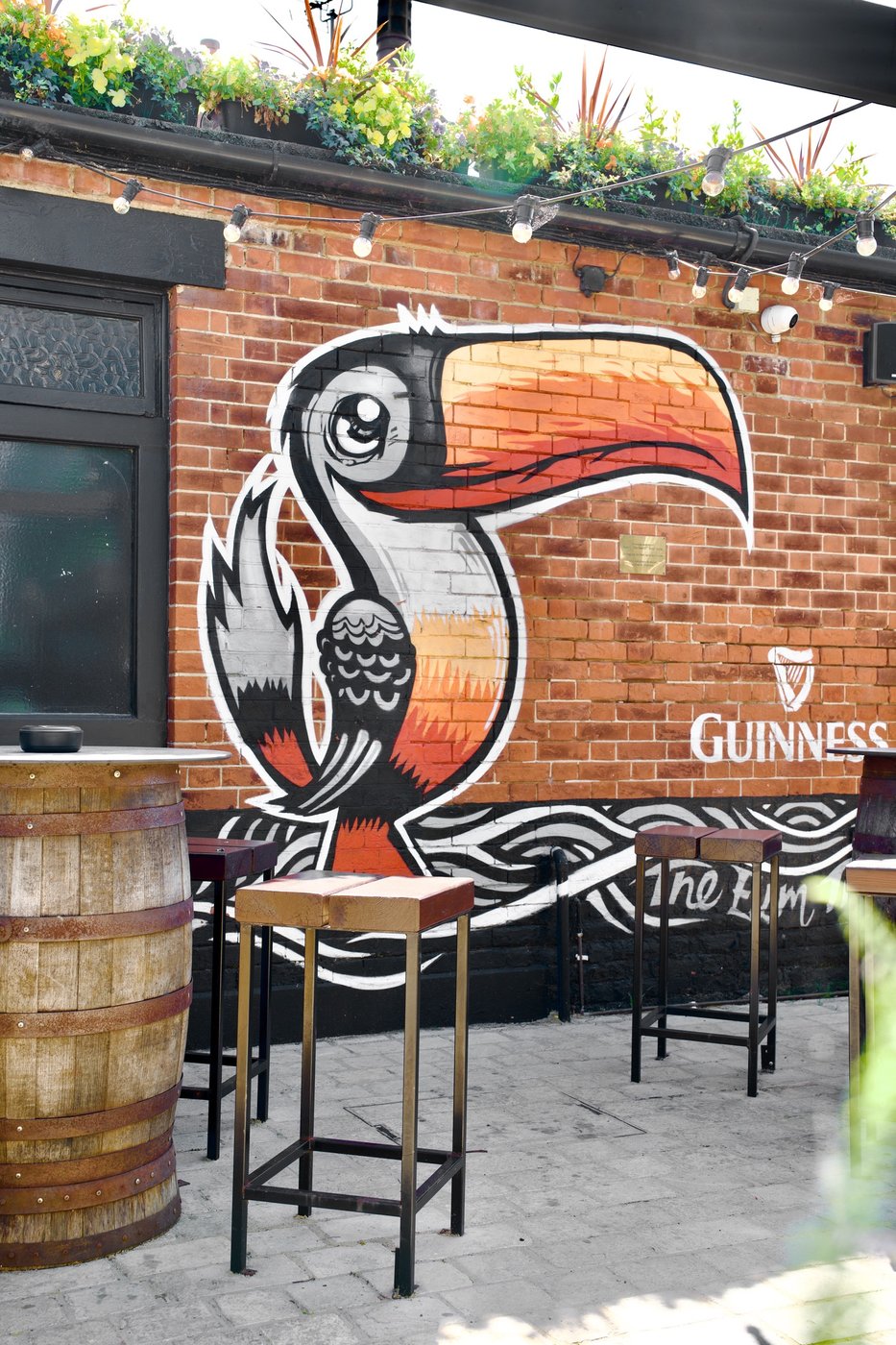 A painted toucan Guinness mural on the brick garden wall, barrel tables in front