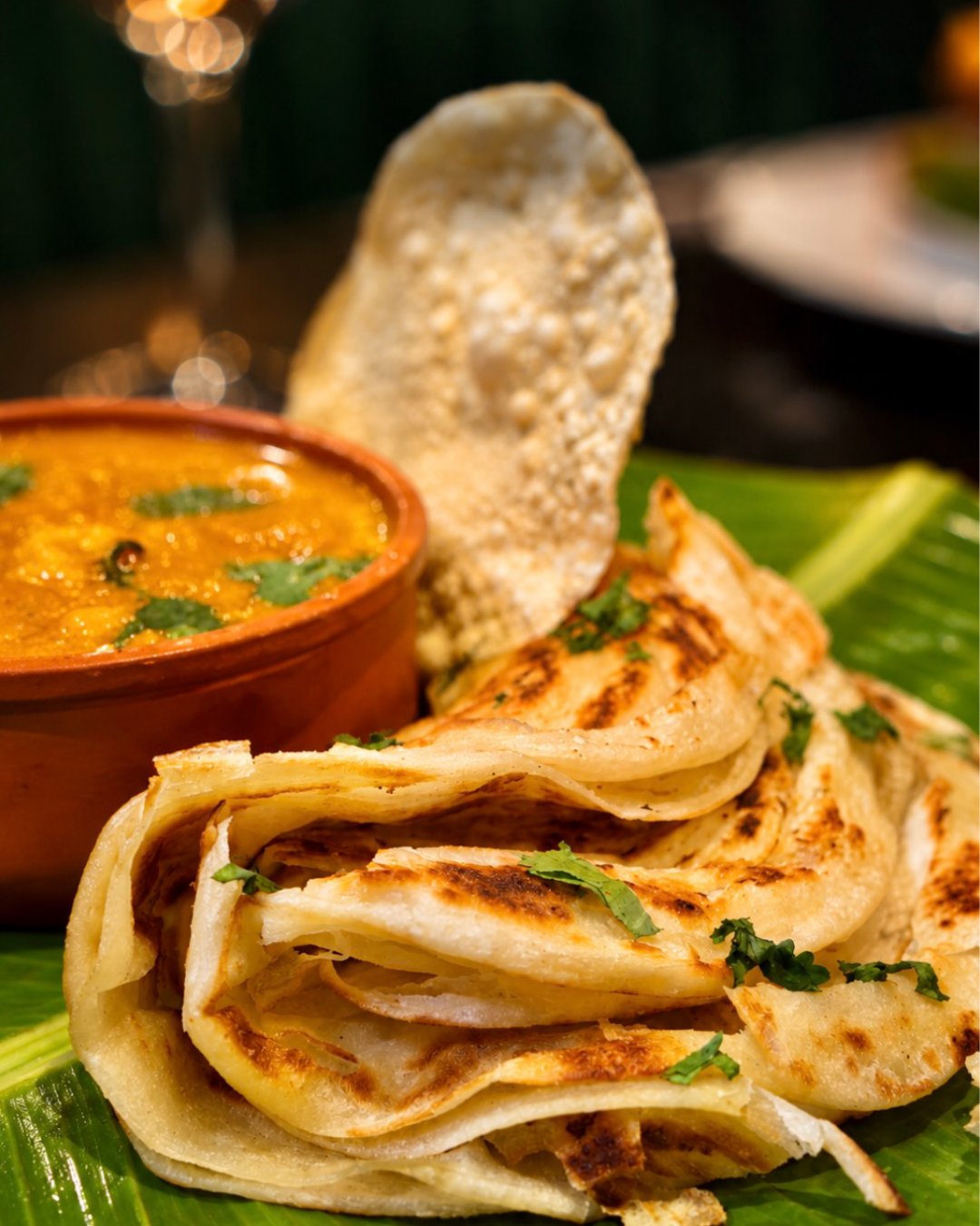 A terracotta bowl of curry served with folded paratha and a poppadom on a banana leaf