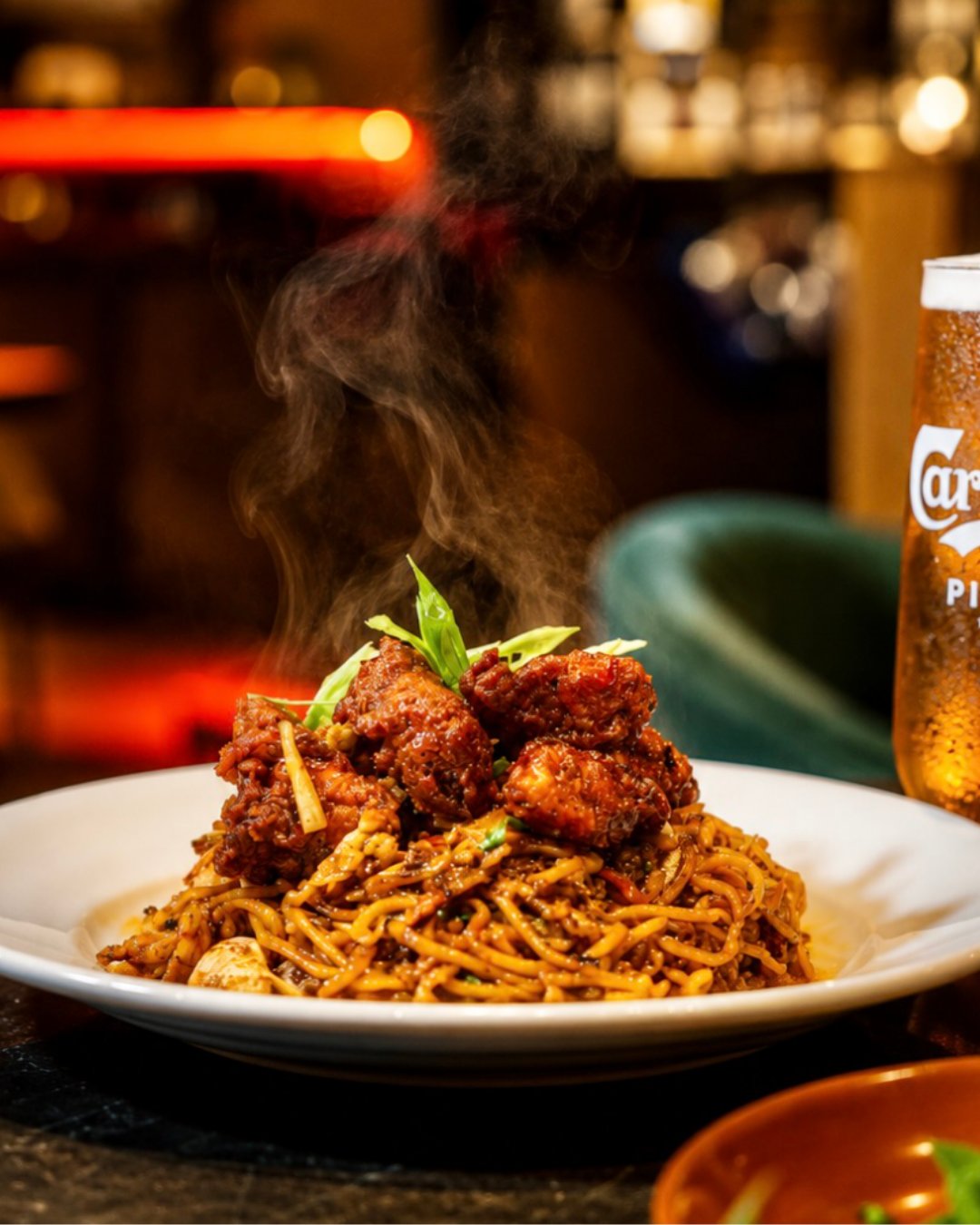 A bowl of Sri Lankan noodles topped with fried chicken, steaming, a lager behind