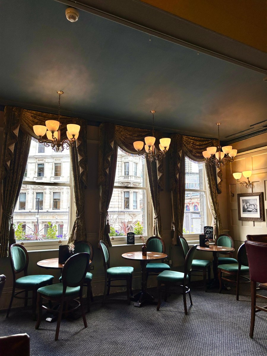 A room of round wooden tables and green velvet chairs under brass chandeliers, tall curtained windows looking onto the street