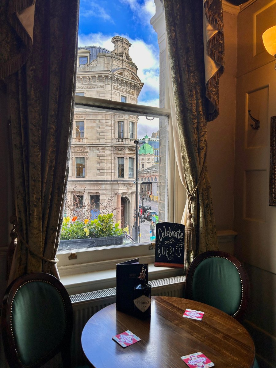 A round table for two by a tall curtained window, looking out across the street to a stone building with a green copper dome