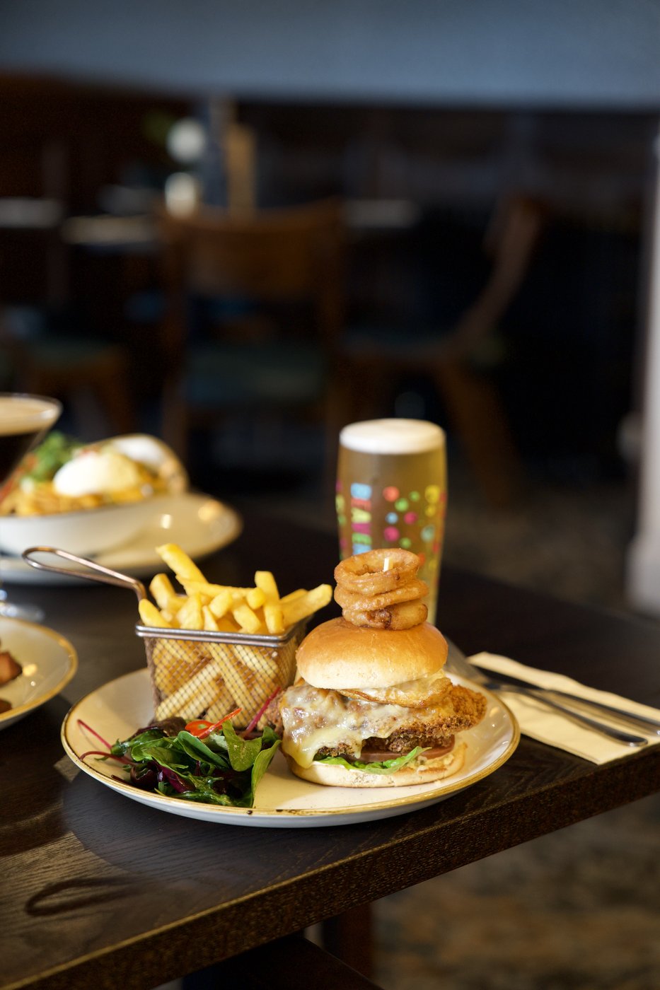 A burger topped with onion rings, with a basket of fries and dressed leaves, a pint on the table behind