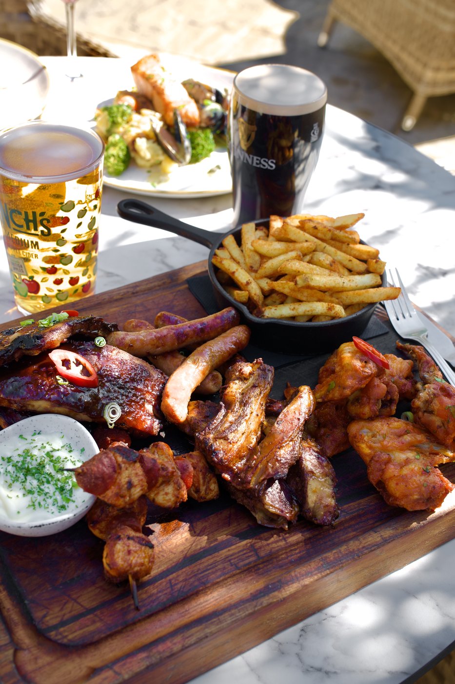 A sharing board of grilled meats, skewers and wings with a cast-iron basket of fries and a dip, alongside a Guinness and a lager