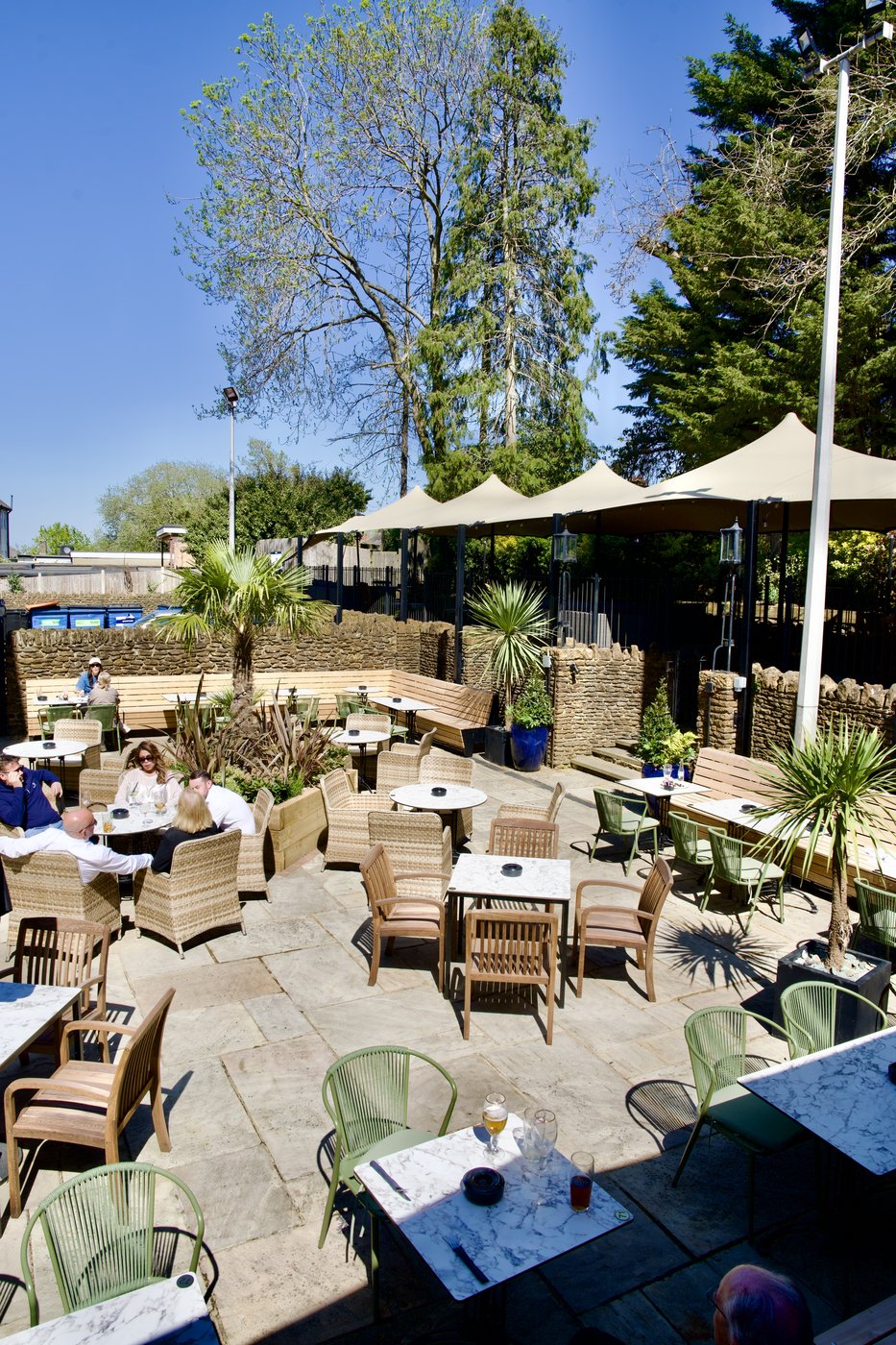 The garden terrace: rattan chairs and tables under parasols, with palms and planting, under a blue sky