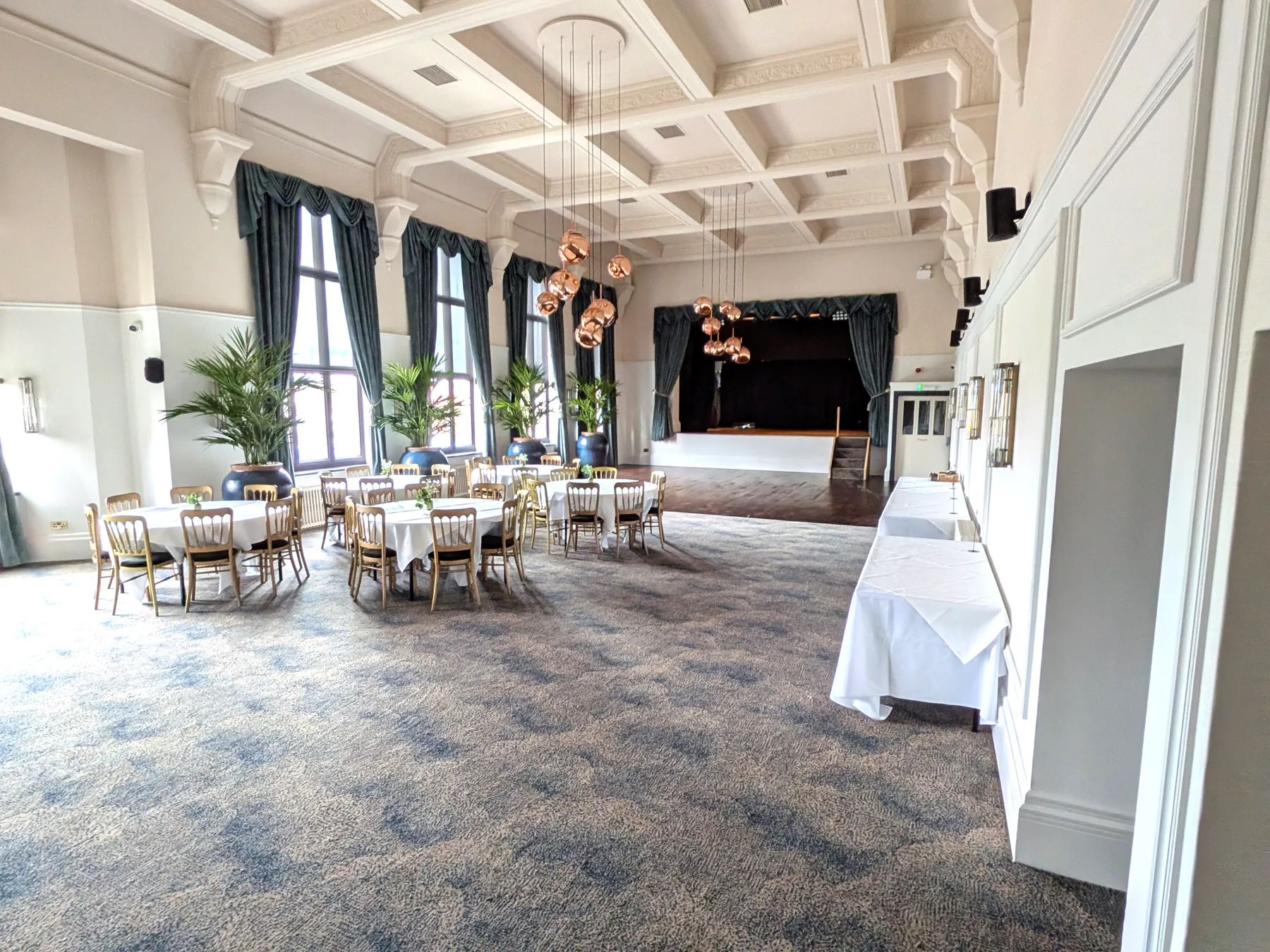 The Grand Suite laid for a function: round tables and gold chairs under clusters of copper pendant lights, with the stage and its green curtains beyond
