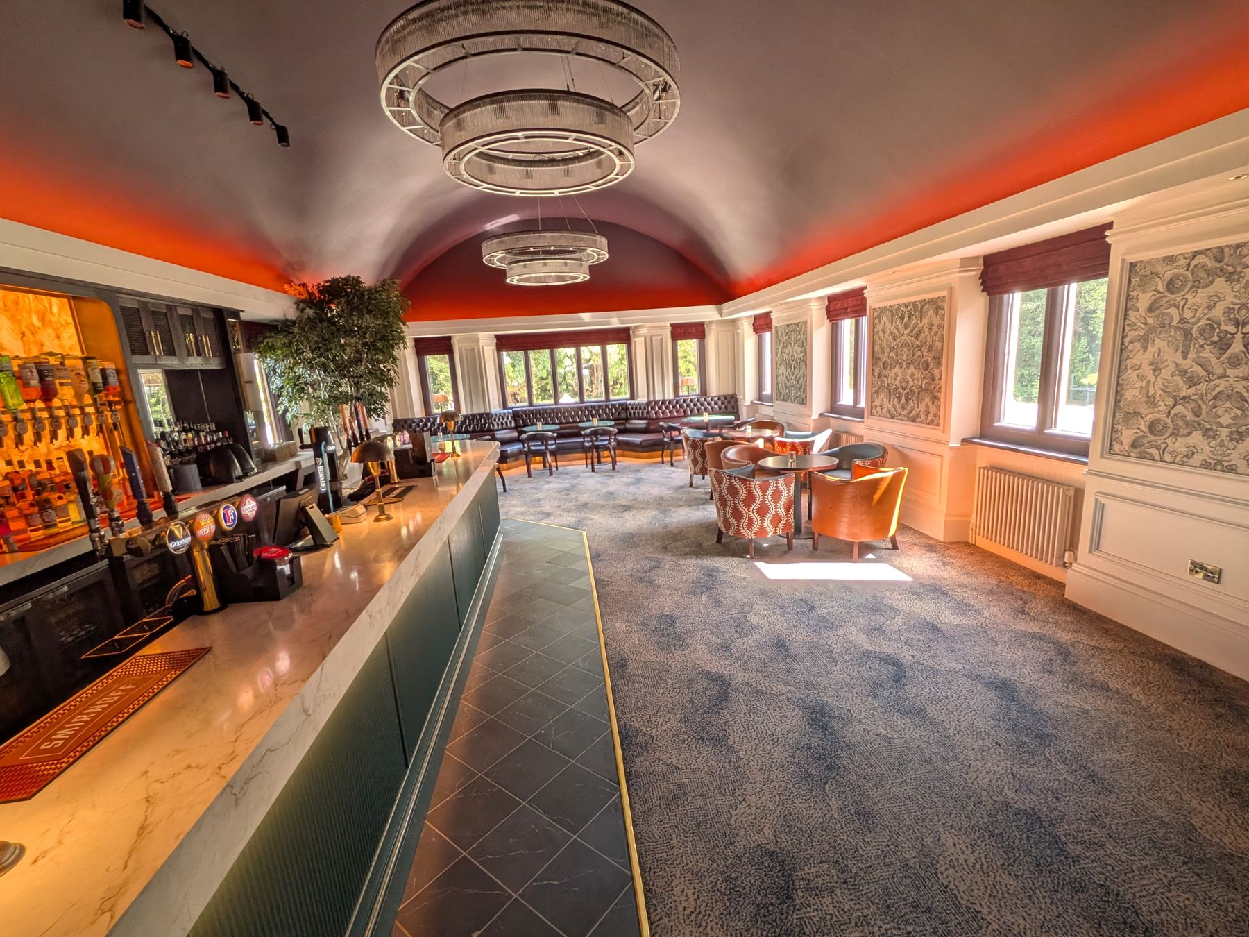 The private bar: a marble-topped bar with the taps along it, ring chandeliers under a barrel ceiling, and leather banquettes and tub chairs by the windows