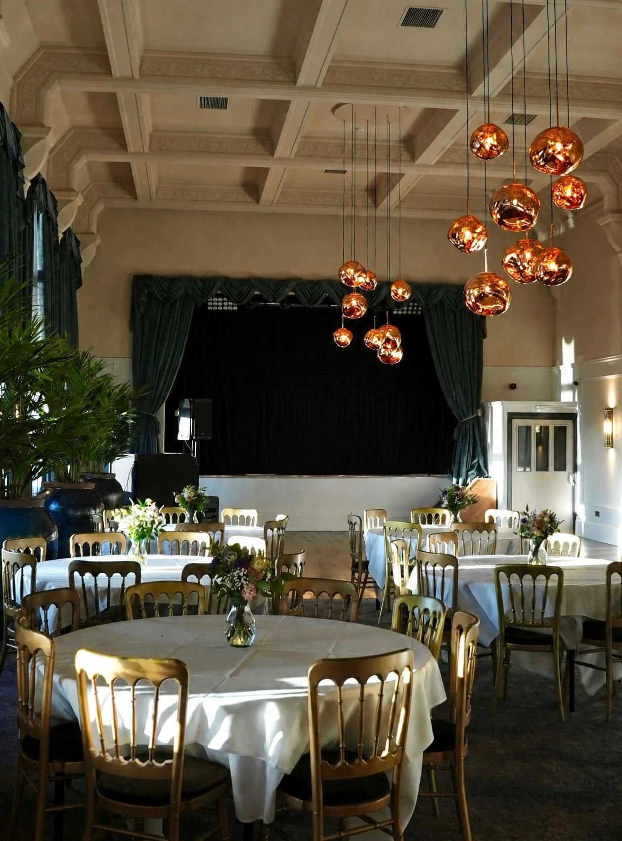 Round tables in white linen, each with a vase of flowers, under clusters of copper pendant lights in the Grand Suite
