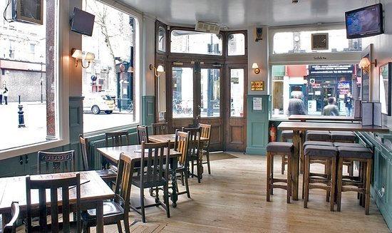 Wooden tables and chairs and high stools in a green-panelled corner room, tall windows looking onto the street