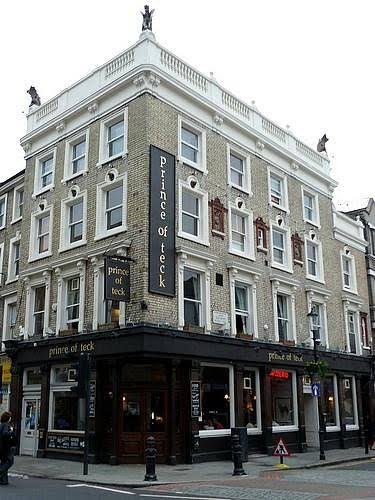The Prince of Teck on its corner of Earls Court Road: white stucco upper floors with stone figures on the parapet above a black pub front and hanging sign