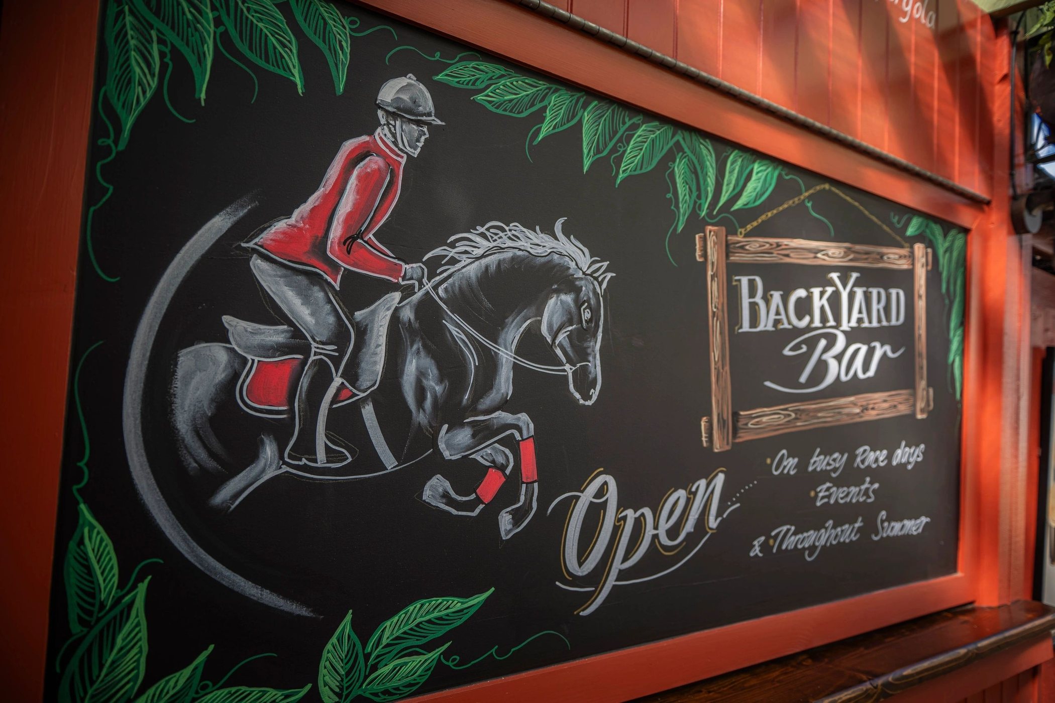 A hand-painted Backyard Bar sign: a jockey and racehorse chalked in white on black, with the bar's opening times beside it