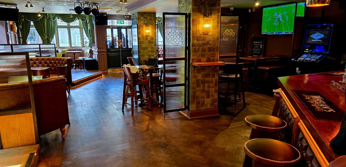 A screen showing sport above the bar at Whelan's Uxbridge, with green curtains and stained-glass windows behind